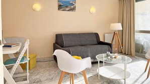 A modern hotel room with a sofa, a small round table, a white chair, a glass coffee table, and a painting on the wall.