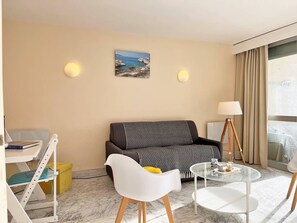 Una habitación de hotel moderna con un sofá, una pequeña mesa redonda, una silla blanca, una mesa de centro de vidrio y un cuadro en la pared.