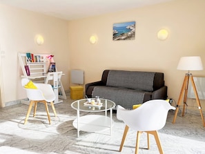 En stue med en sofa, to hvide lænestole, et sofabord, en reol, en lampe og et maleri af et kystlandskab.
