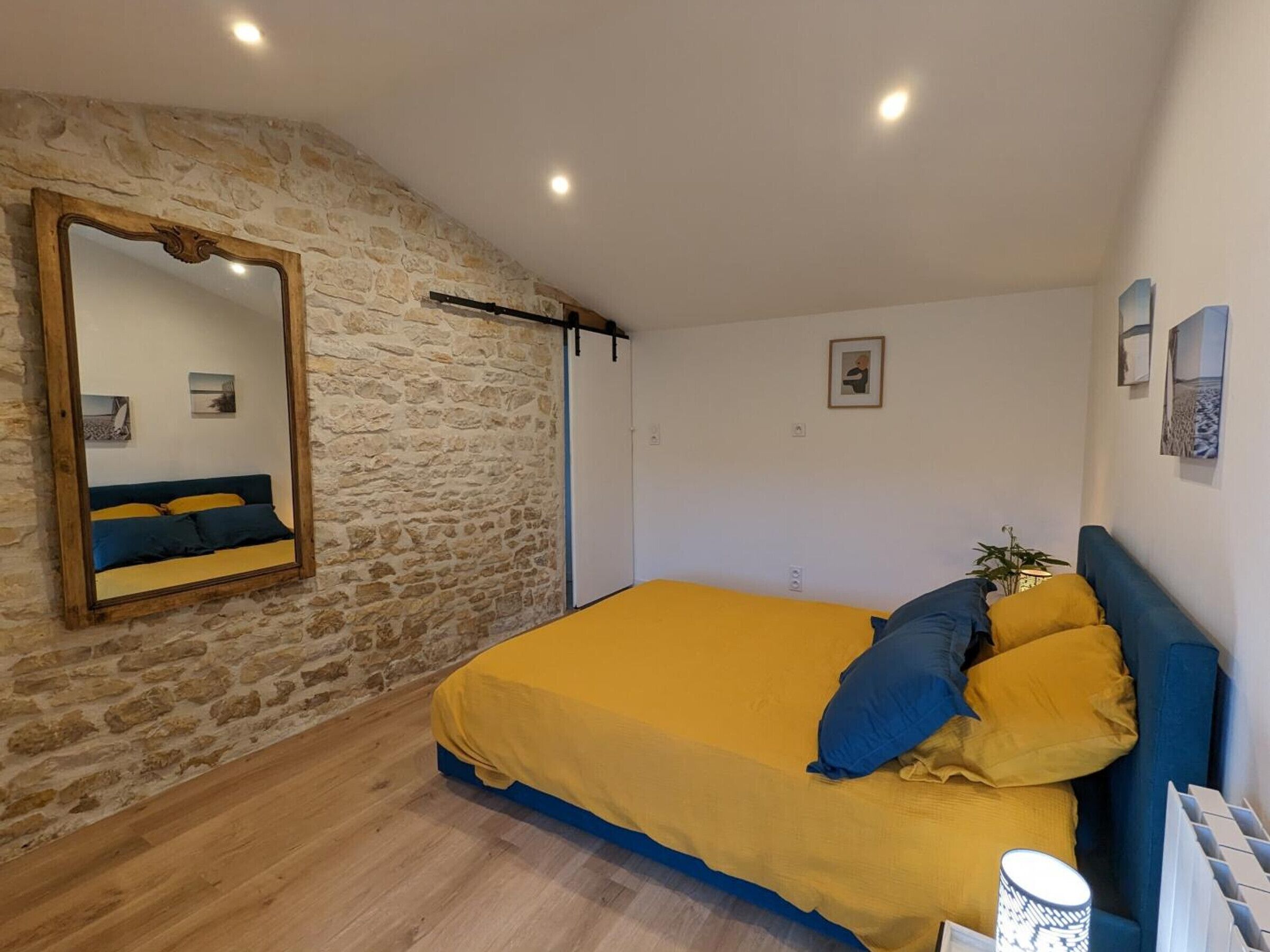 Une chambre avec un mur en pierre, un lit avec une literie jaune et bleue, un miroir à cadre en bois et une table de chevet avec une lampe.