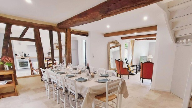 A dining area with a long table set for six, surrounded by white chairs, with exposed wooden beams on the ceiling and a kitchen in the background. - Family Retreat with Garden, Near Leisure Activities (Saint-Poix)