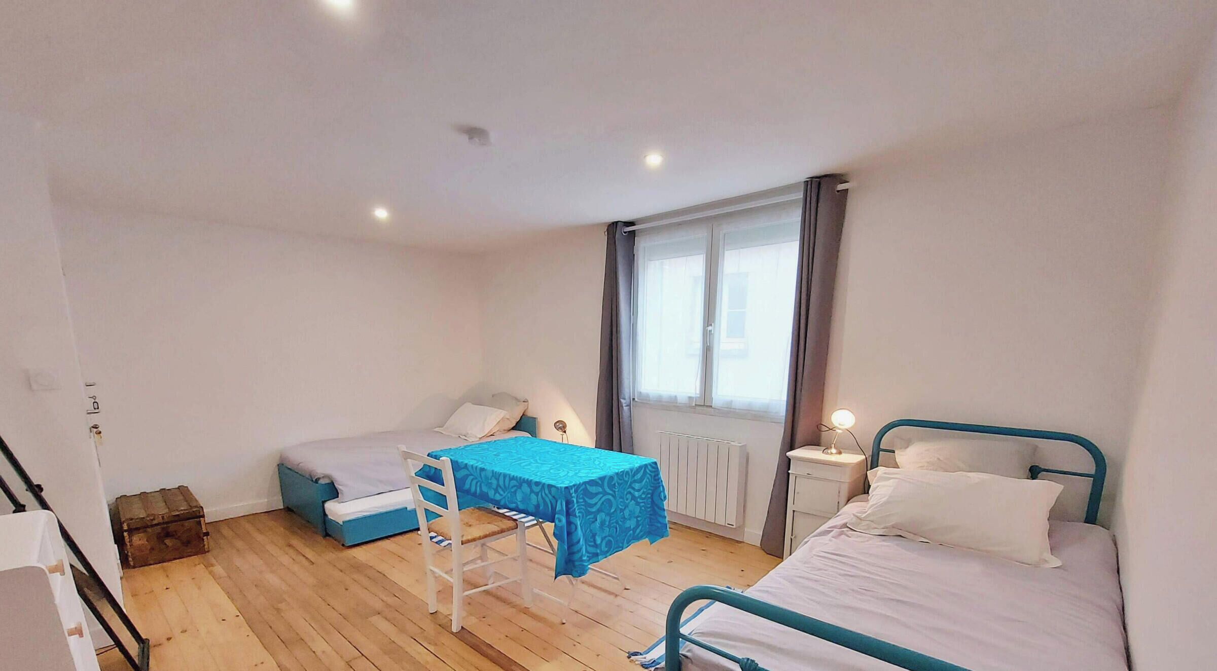 Una habitación pequeña y bien iluminada con dos camas, una mesa y una silla. Cuenta con una ventana con cortinas, un radiador y un mueble pequeño.