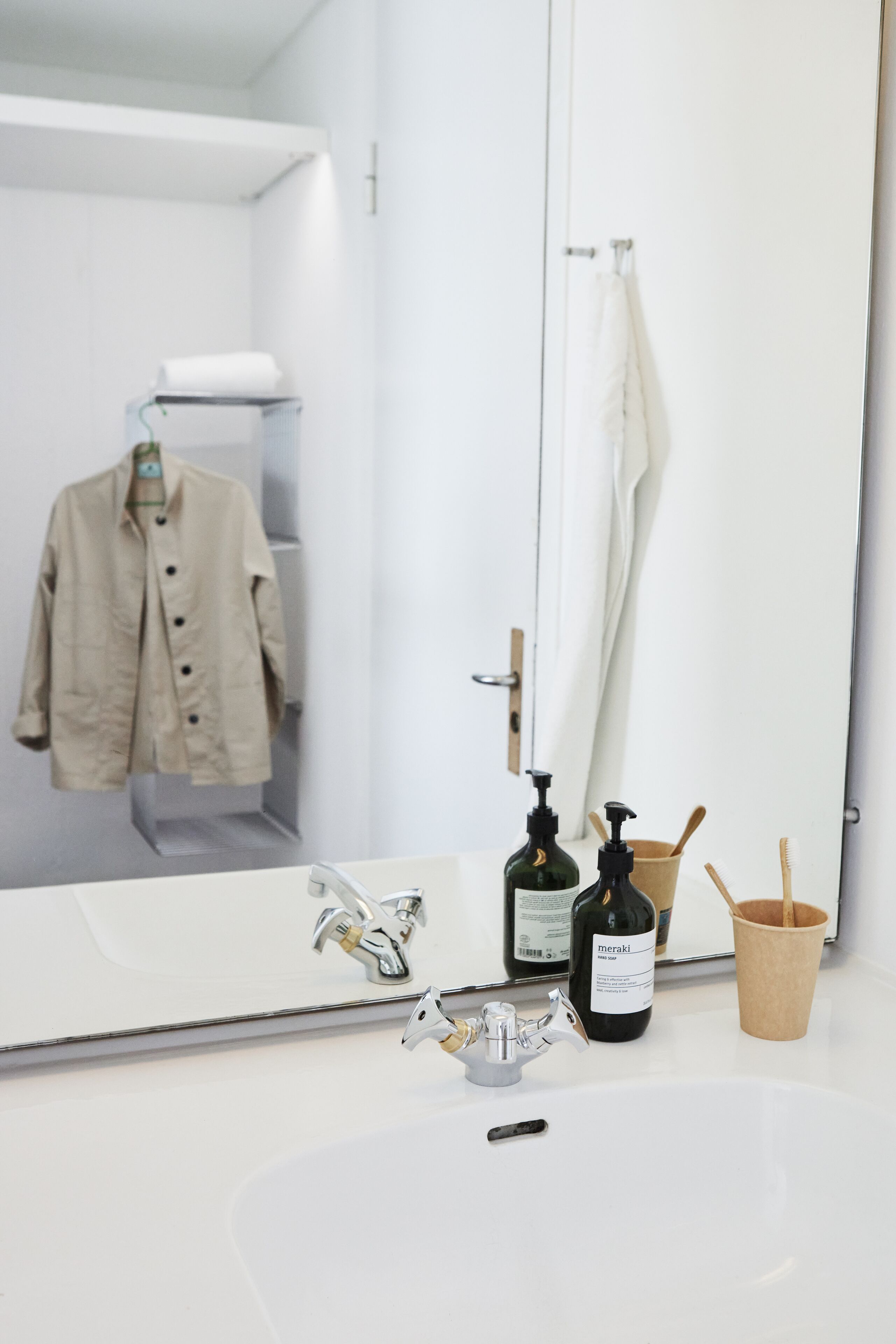 A bathroom with a white sink, a mirror, a bottle of Meraki soap, a toothbrush holder, and a hanging towel.