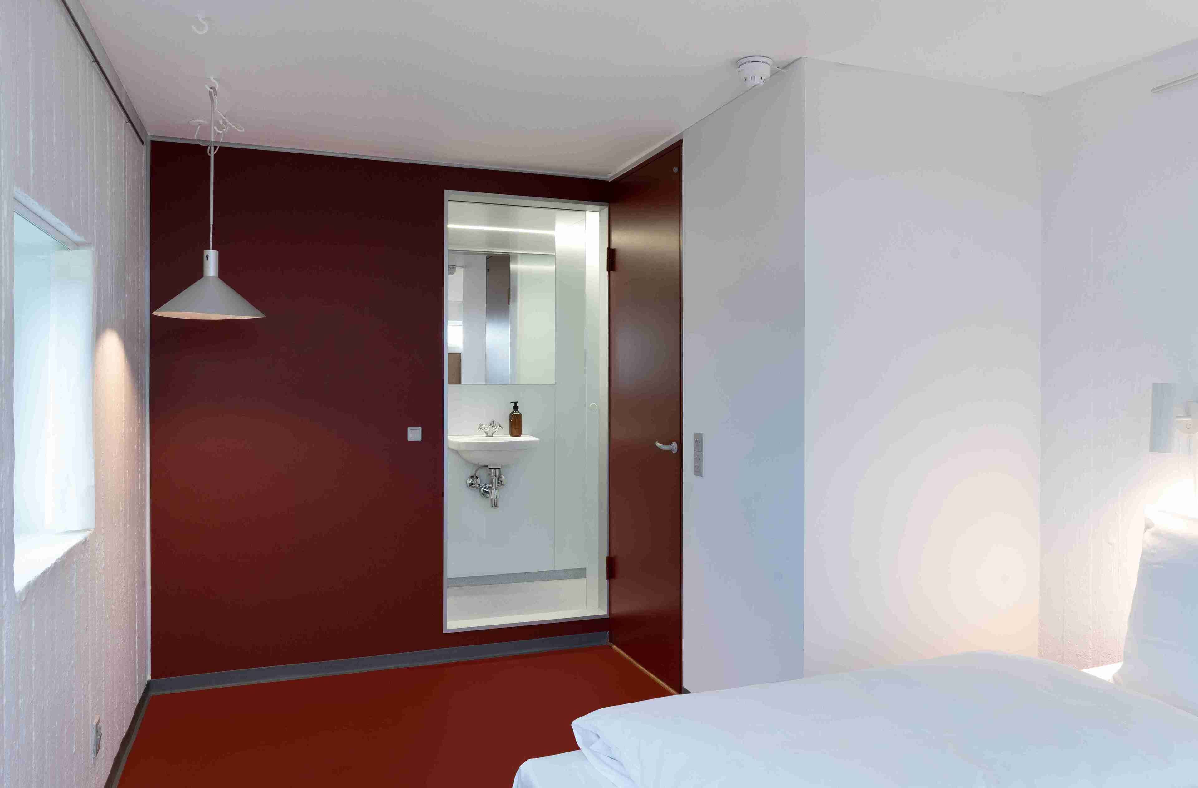 A hotel room with a bed, a bedside table, a hanging light fixture, a window, and a door leading to a bathroom with a mirror and sink.