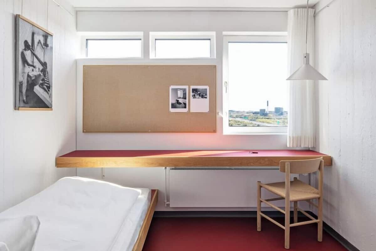 A hotel room with a bed, a desk with a chair, a bulletin board, and a window with a city view.