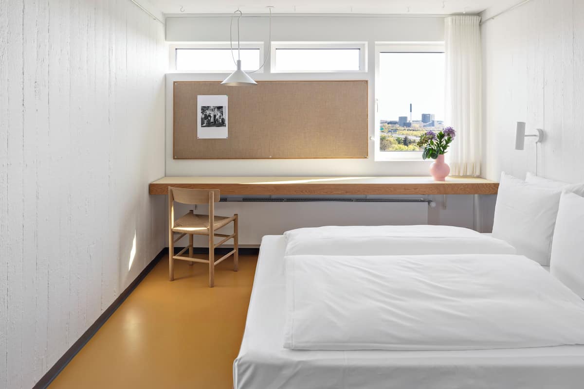 A bedroom with a bed, a desk, a chair, a window with a city view, and a wall-mounted light fixture.