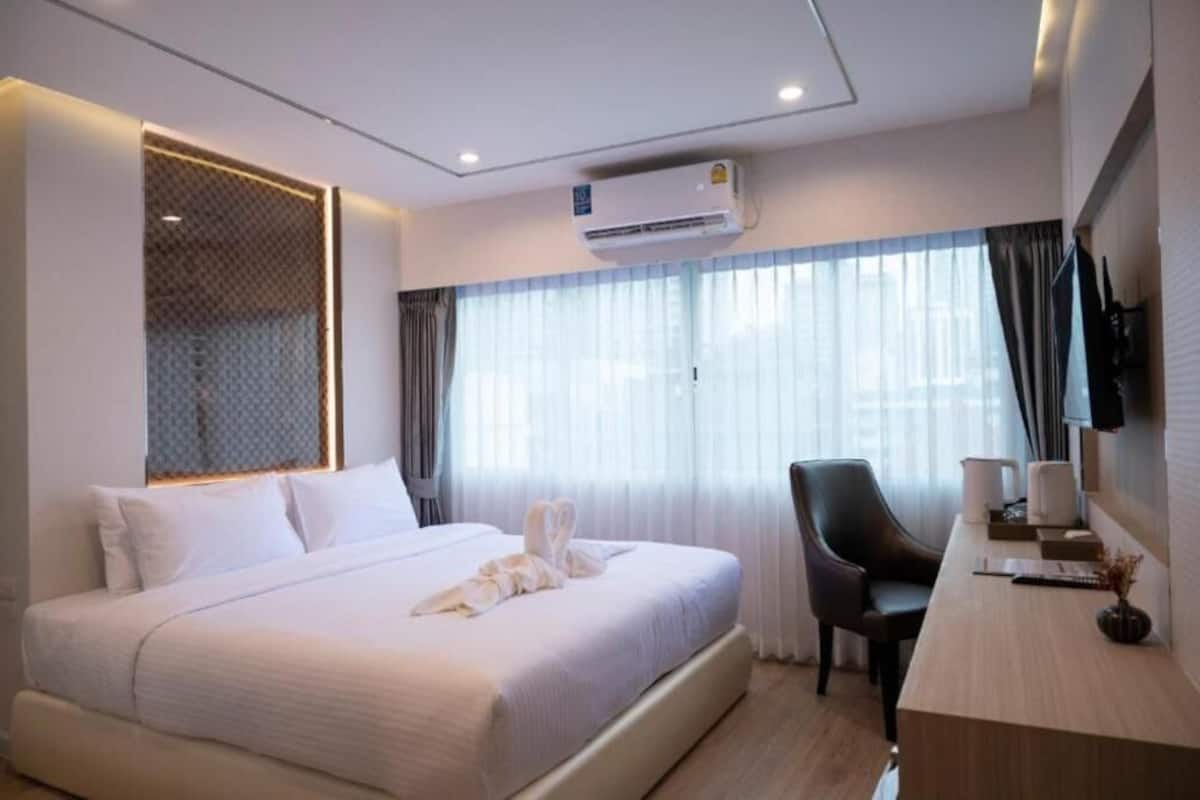 A hotel room with a bed, a desk, a chair, an air conditioning unit, and a large window with curtains.