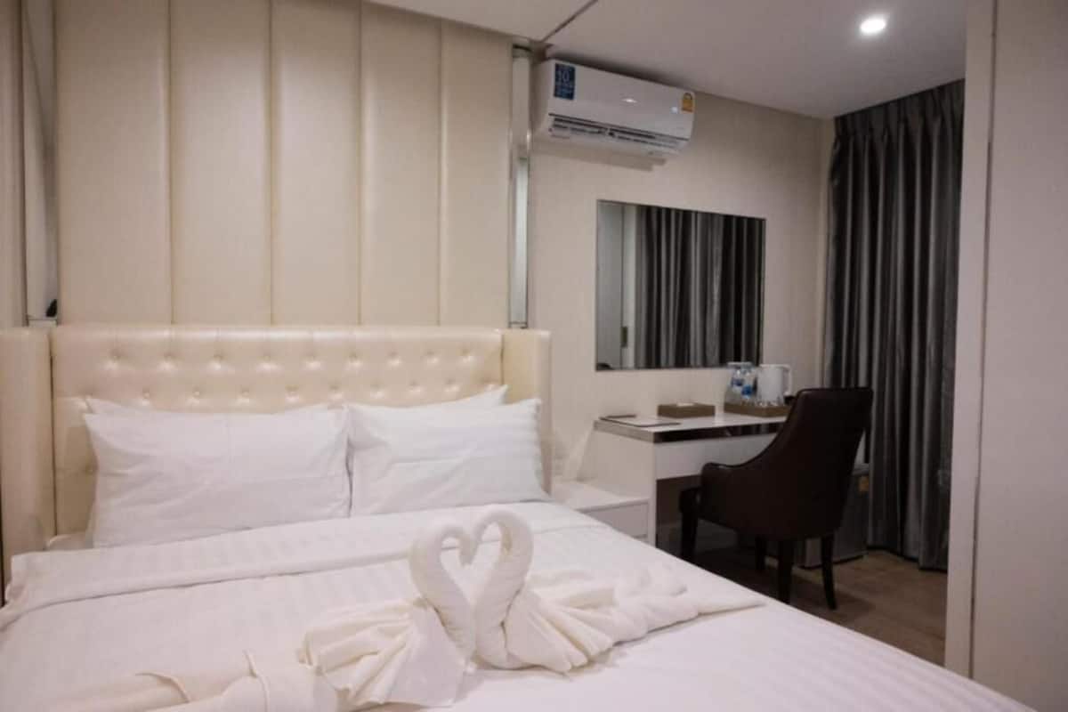 A hotel room with a bed, a desk, a chair, a mirror, and an air conditioning unit.