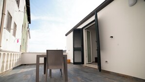 A balcony with a wicker chair and table, a white building with shutters, and a clear sky.