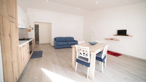 A modern kitchen with wooden cabinets, a dining area with a table and chairs, a blue sofa, and a TV on a shelf.