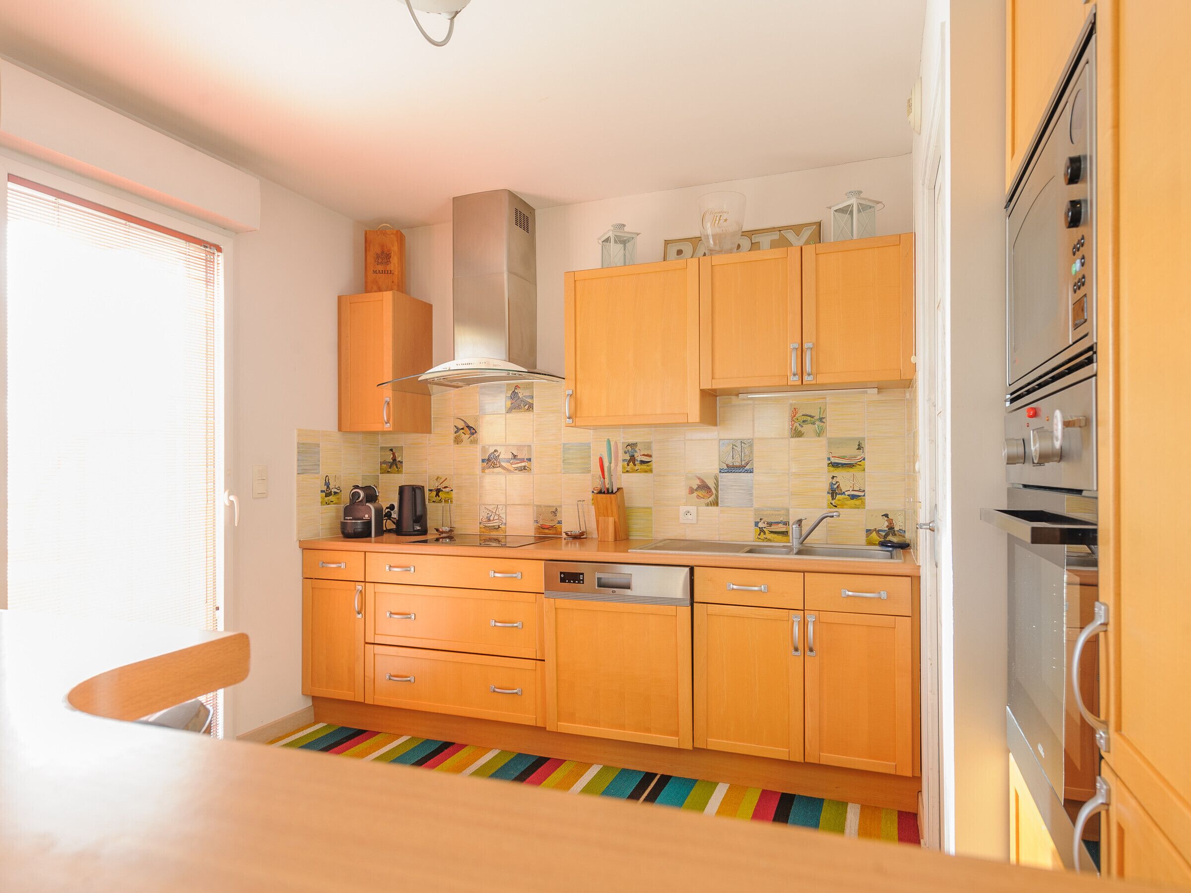 Una cocina con armarios de madera, extractor de acero inoxidable, salpicadero de azulejos con motivos náuticos, alfombra a rayas de colores y una ventana con persianas.
