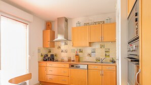 Eine Küche mit Holzausstattung, einer Edelstahl-Dunstabzugshaube, einer gefliesten Spritzschutzwand mit maritimer Kunst, einem farbenfrohen Streifen Teppich und einem Fenster mit Jalousien.