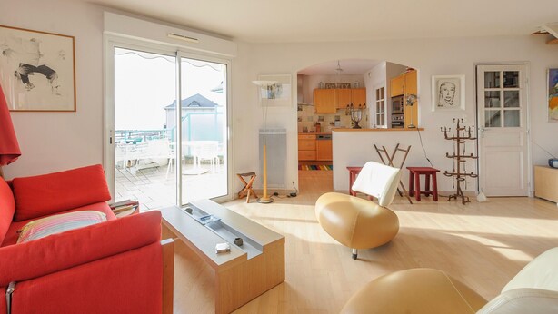 Ein modernes Wohnzimmer mit einem roten Sofa, einem beigefarbenen Sessel und einem Couchtisch. Eine Schiebetür aus Glas führt auf einen Balkon mit Gartenmöbeln. Im Hintergrund ist eine Küche mit Holzschränken und einer weißen Arbeitsplatte zu sehen.