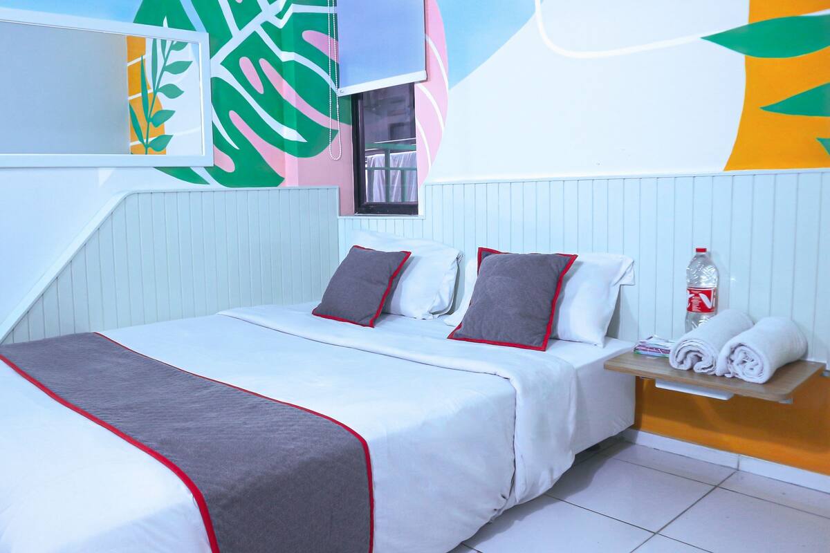 A neatly made bed with a grey and white bedspread, two pillows, and a red-lined blanket. A bottle of water and rolled towels are on a wooden side table next to the bed.