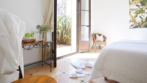 Un dormitorio con una cama, una silla de mimbre, una mesita con una planta y vistas al exterior.