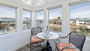 Une pièce avec une table ronde et deux chaises, de grandes fenêtres donnant sur un port de plaisance et des bâtiments, et un objet décoratif posé sur la table.