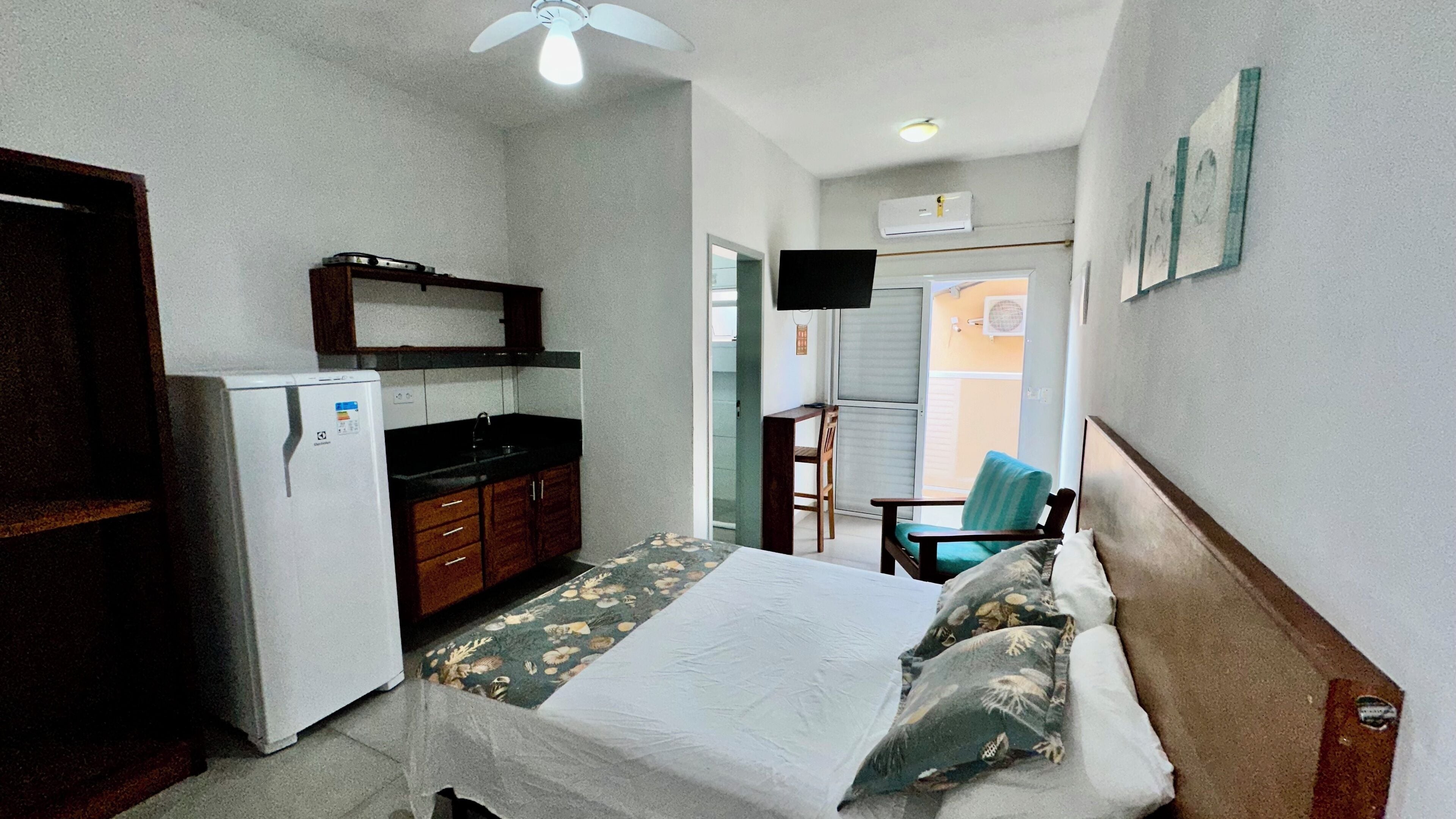 A compact hotel room with a bed, a small kitchenette, a ceiling fan, and a TV.