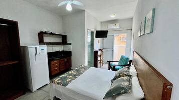 Quarto de hotel compacto com cama, pequeno espaço de cozinha, ventoinha no teto e televisão.
