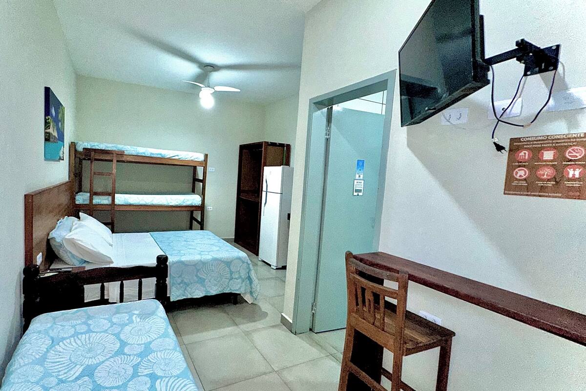 Une petite pièce compacte comprenant deux lits superposés, un lit simple, un bureau, une chaise, un réfrigérateur, un ventilateur de plafond et un téléviseur fixé au mur.