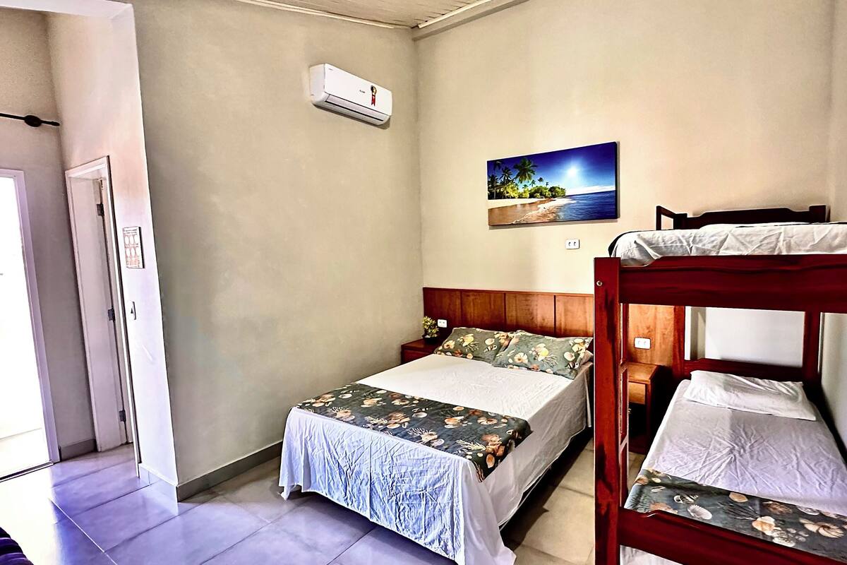 Une chambre avec un lit superposé, un lit simple avec une literie à motifs floraux, une table de chevet avec une plante et un tableau représentant une scène de plage.