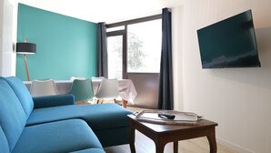 A living room with a teal wall, a large sofa, a dining table with chairs, a TV mounted on the wall, and a small wooden table with a tray. - Beautiful 3-room apt, renovated, central, facing slopes, 6 pers., balcony, parking (Pra-Loup)