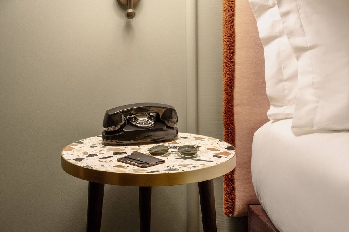 A neatly arranged hotel room with a round table, a telephone, a pair of sunglasses, and a lamp.