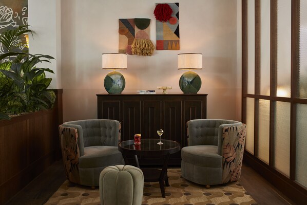 A lounge area with two armchairs, a round table, and two lamps. There is a plant to the left and a window with blinds to the right. - The Hoxton, Berlin (Berlin)