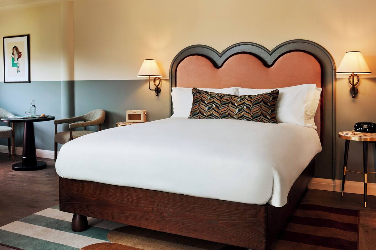 A hotel room with a large bed, two bedside tables, a chair, a small table, and a wall-mounted lamp.