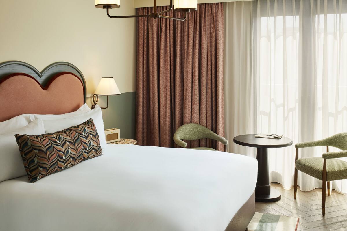 A hotel room with a large bed, two chairs, a small table, a lamp, and a view of the outdoors through curtains.