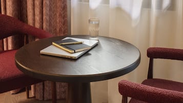 Une table ronde avec un carnet, un stylo et un verre d’eau, entourée de chaises à revêtement rouge, dans une pièce dotée de rideaux et d’un plancher en bois.