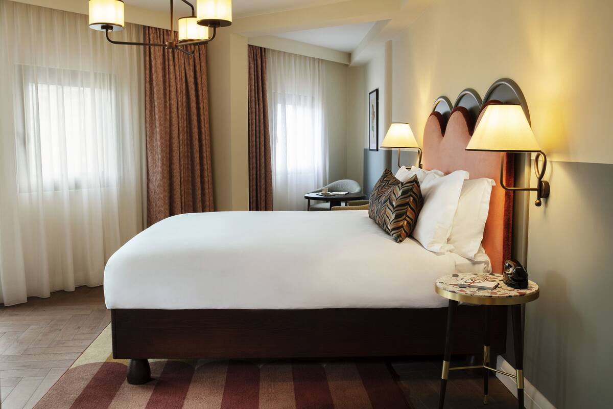 A hotel room with a large bed, a bedside table with a lamp, a chair, and a desk with a telephone.