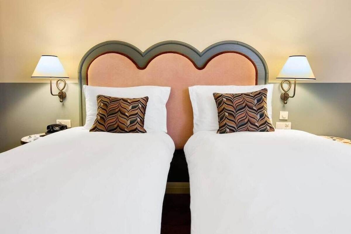 A hotel room with two beds, each with a white bedspread and patterned pillows, flanked by wall-mounted lamps.