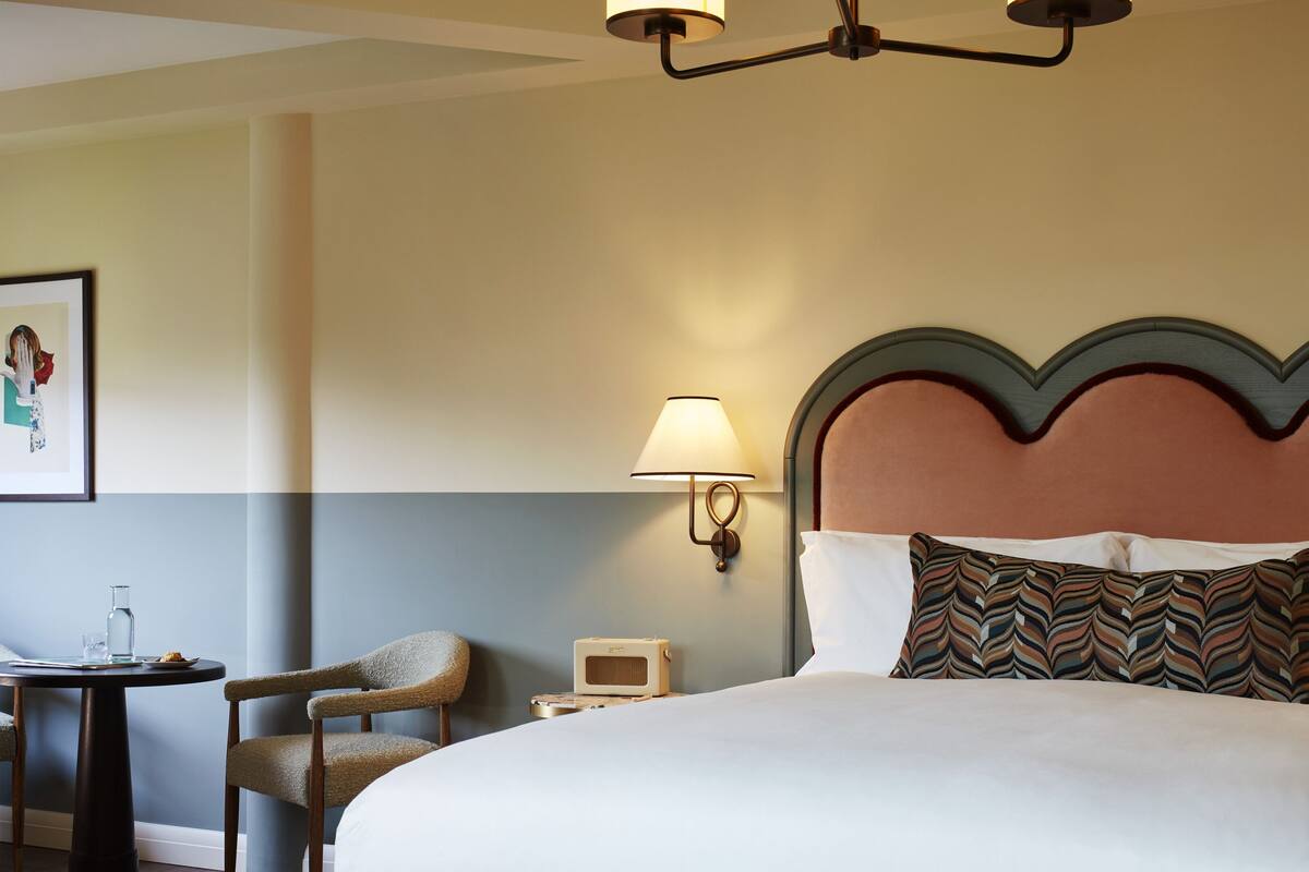 A hotel room with a large bed, a bedside table with a lamp, a chair, and a small table with a glass and a book.