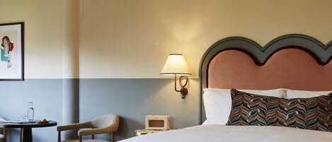 A hotel room with a large bed, a bedside table with a lamp, a chair, and a small table with a glass and a book.