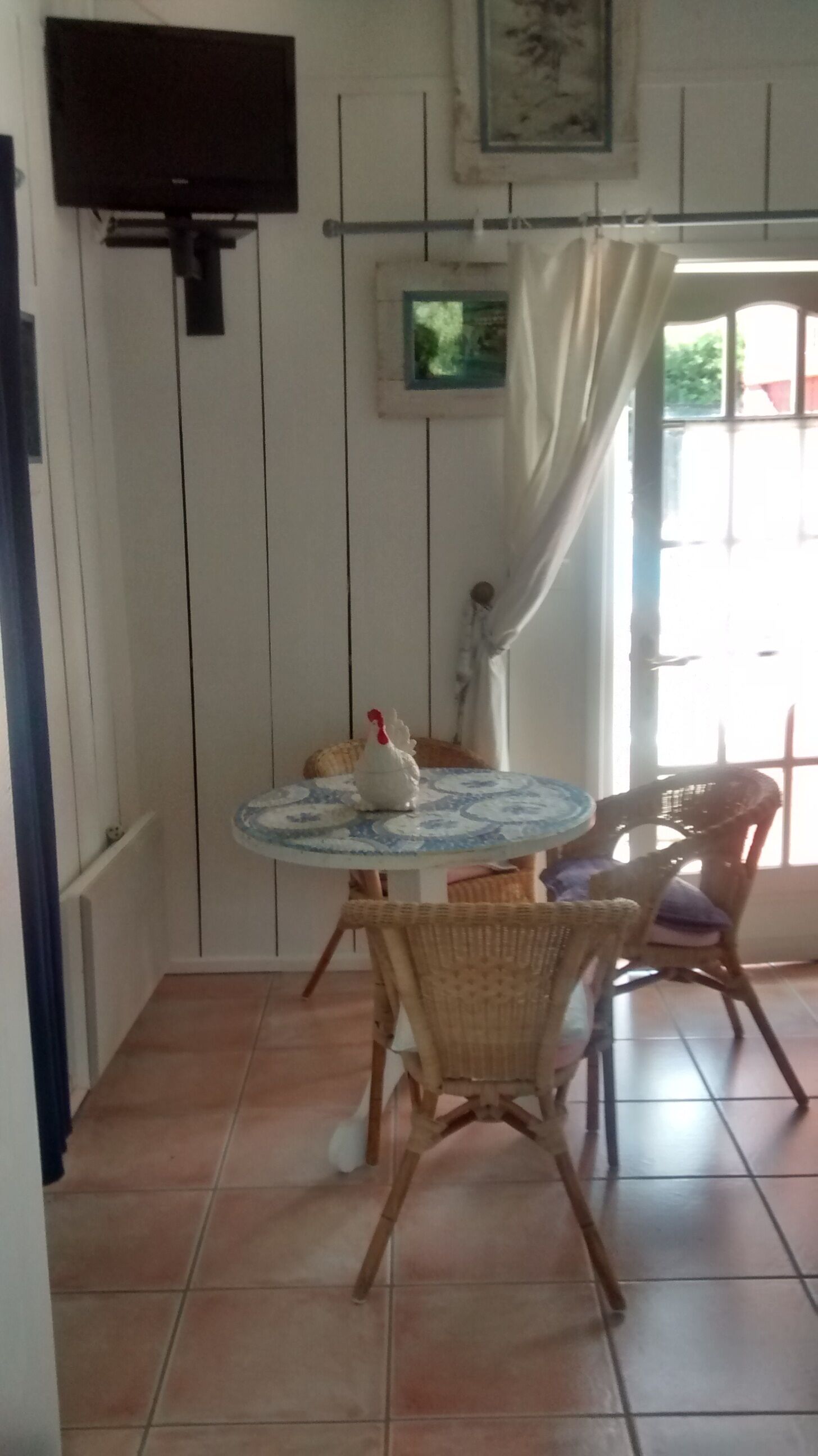 Uma área de refeições com uma mesa redonda, cadeiras de vime, um pequeno galo decorativo e uma televisão na parede.