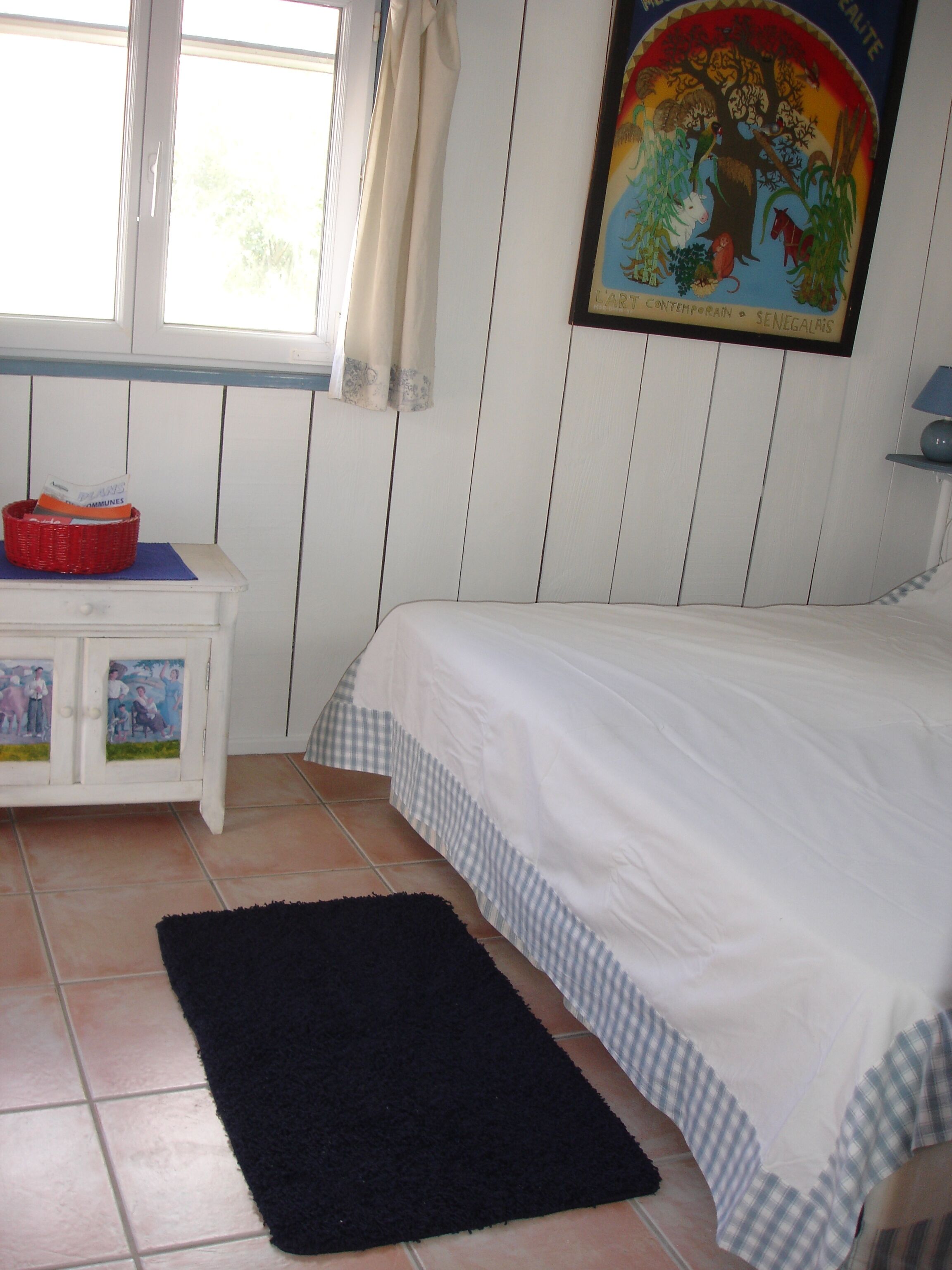 A bedroom with a bed, a nightstand, a window with curtains, a painting on the wall, and a black rug on the floor.
