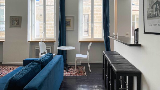 Ein modernes Wohnzimmer mit einem blauen Sofa, einem weißen Sessel, einem kleinen runden Tisch und einer schwarzen Bank. Zwei Fenster mit Vorhängen, ein gerahmtes Bild an der Wand und ein gerahmtes Foto rechts.