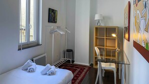 Ein Hotelzimmer mit Bett, Fenster, Kleiderständer, Schreibtisch mit Stuhl und Wand mit Kunstwerk.