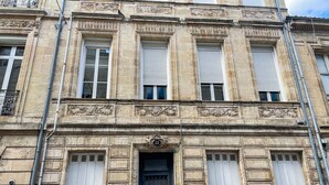 Ein historisches Gebäude mit aufwendiger Steinmetzarbeit, großen Fenstern mit Fensterläden und einem zentralen Eingang mit einer Tür.