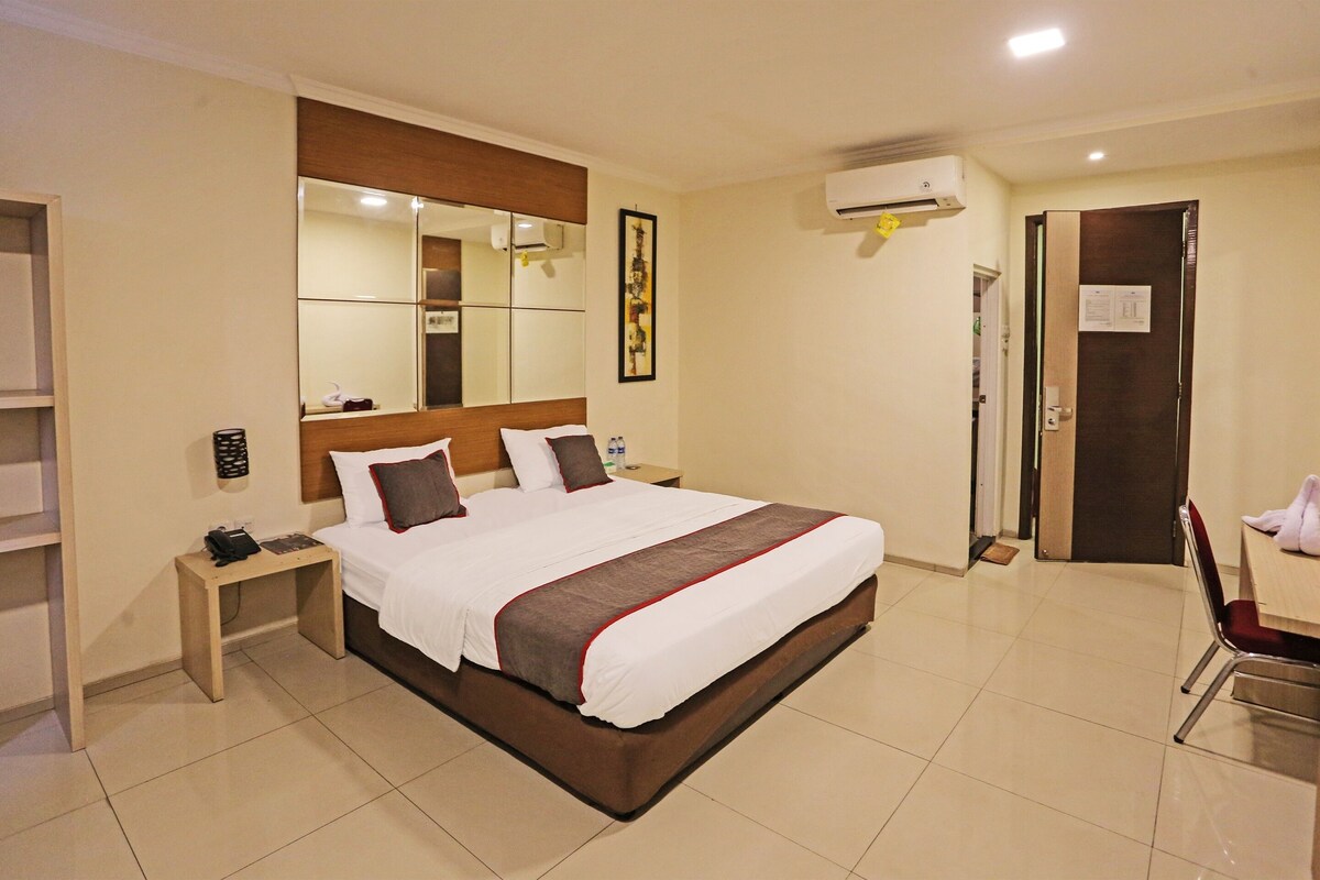 A hotel room with a large bed, bedside table, and a mirror. There is a door to the right and a shower area.