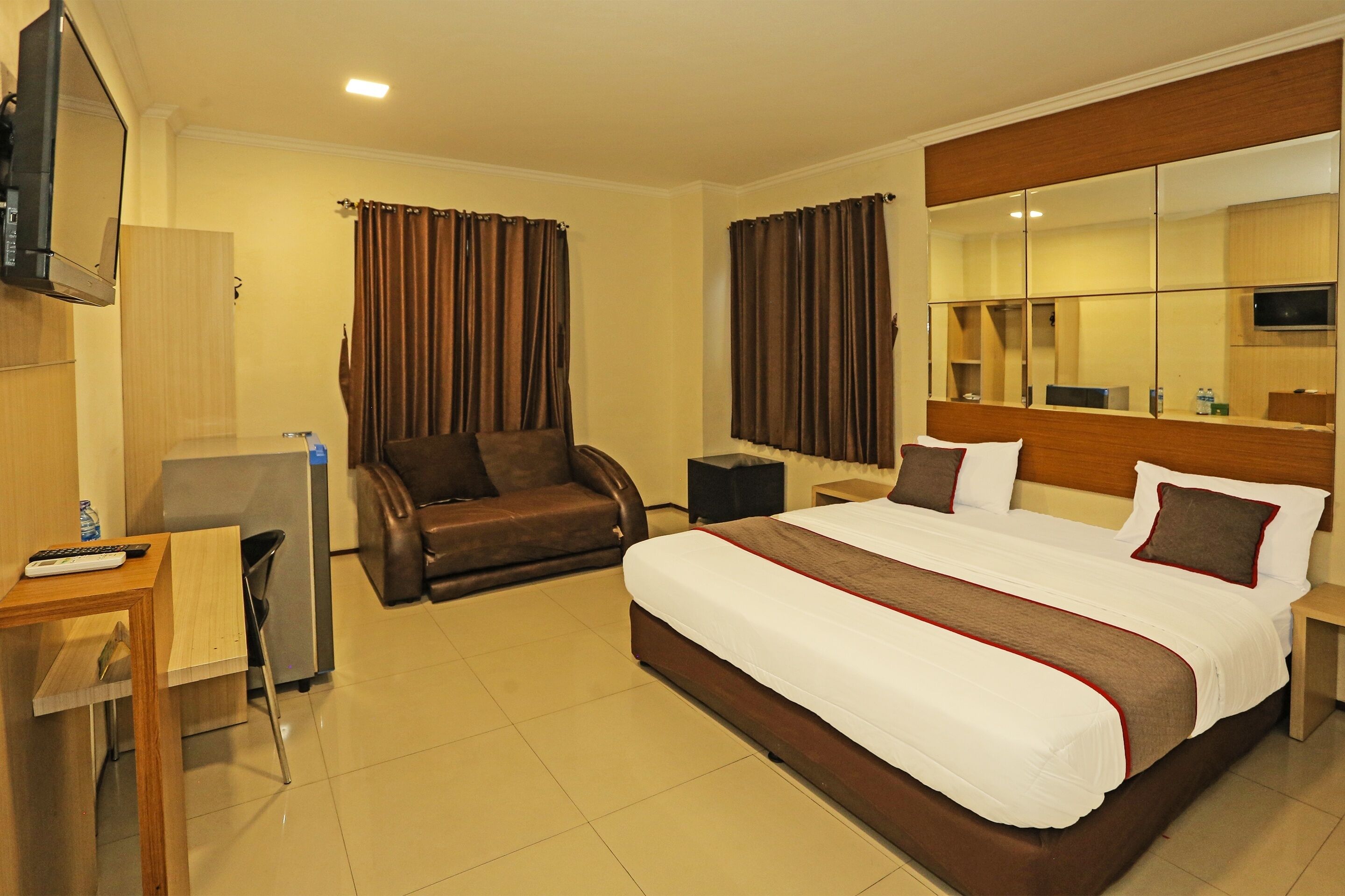 A hotel room with a large bed, a desk with a chair, a TV mounted on the wall, and a mirror above the bed.