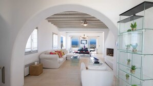 A spacious living area with a white sofa, a glass coffee table, and a glass shelving unit with plants. - 360 Sunset Villa with Private Pool (Mykonos)