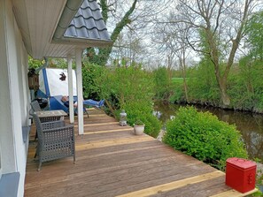 A covered outdoor area with a wooden deck, seating, and a view of a water body surrounded by greenery.