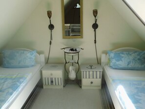 A small, triangular room with two beds, each with a floral-patterned pillow. There is a small table with a vase in the center, flanked by two wall-mounted lamps.
