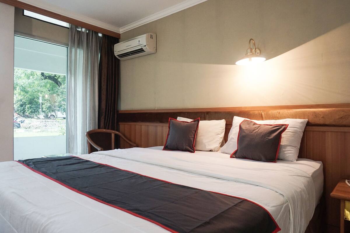 Ein Hotelzimmer mit einem großen Bett, einem Fenster mit Vorhängen, einer Klimaanlage, einer Nachttischlampe und einem Holz-Kopfteil.