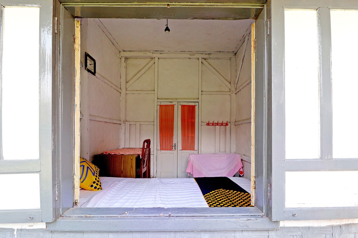A small, single-room accommodation with a bed, a chair, a table, and a window with curtains.