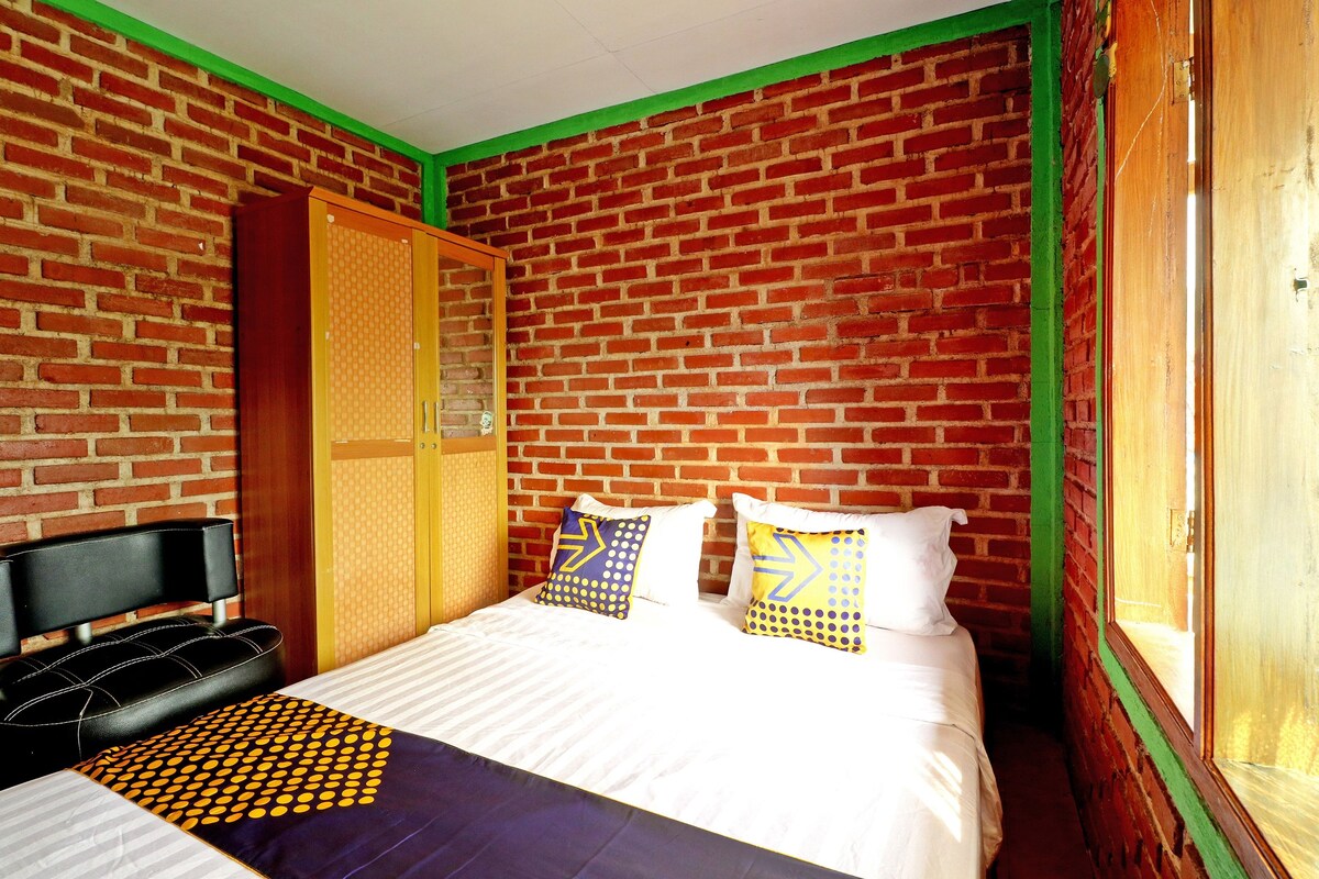 A room with a brick wall, a bed with white bedding and patterned pillows, a wooden wardrobe, and a window with a view.