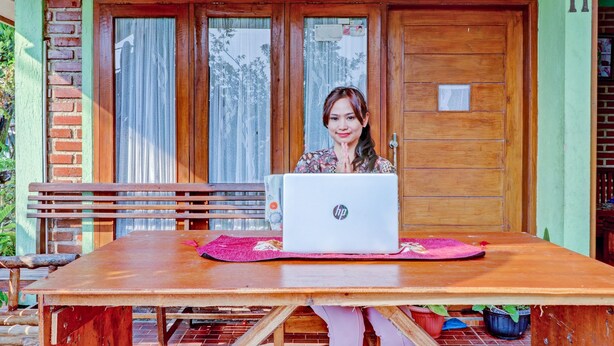 A person sitting at a wooden table with an HP laptop, in front of a wooden door with a window. - OYO Homes 90849 Cibeunying Homestay Cibodas Maribaya (Lembang)
