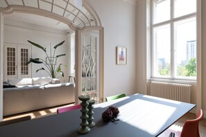 A room with a white table, a green chair, a white sofa, and a large window with a view of buildings and trees. - 2-Bedroom Apartment with Private Library (Wien)