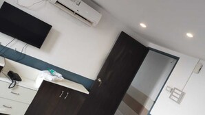 A room with a flat-screen TV mounted on the wall, a desk with a telephone and water bottles, a dark wooden wardrobe, and a door with a handle. - Renuka Executive (Rajgurunagar)